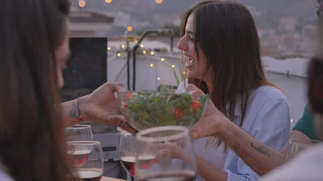 Young Caucasian woman chatting smiling with friends while eating. People gathered celebrating outdoor barbecue. Slow motion happy group having dinner on city rooftop in background. 