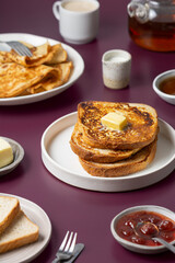 Breakfast food - plates with French toast with butter, crepes, toast bread, jam, honey, tea kettle, coffee cup, milk jug on purple background. Morning meal.