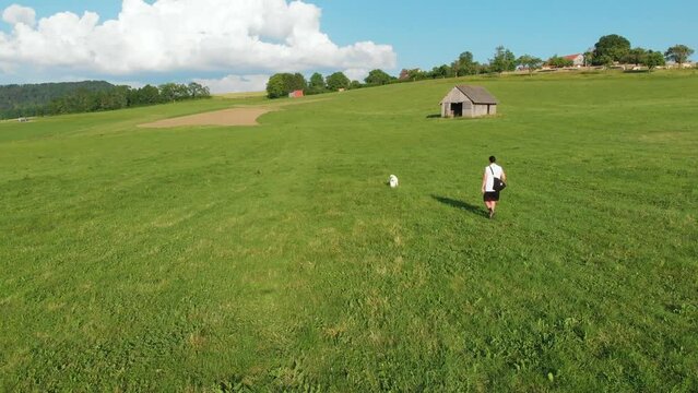 wei&szlig;er Husky Hund und sein Besitzer, ein Junge mit langen Haaren, shorts, einem Achselshirt, einer Umh&auml;ngetasche gehen gemeinsam auf einem Bauernhof Feld auf einer gr&uuml;nen Wiese Gassi im Sommer 4k 