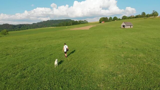 wei&szlig;er Husky Hund und sein Besitzer, ein Junge mit langen Haaren, shorts, einem Achselshirt, einer Umh&auml;ngetasche gehen gemeinsam auf einem Bauernhof Feld auf einer gr&uuml;nen Wiese Gassi im Sommer 4k 