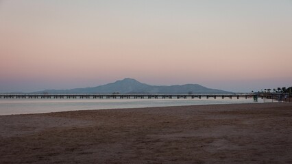 Fototapeta na wymiar Calm sea and the sandy beach with a wooden dock and mountains against the pink sunset