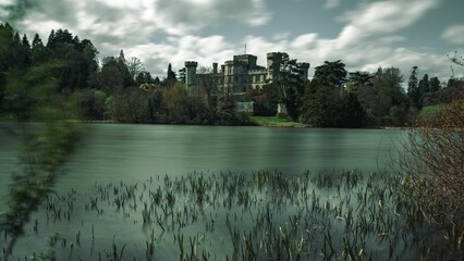 Fototapeta premium Long-exposure waterside view of Eastnor Castle surrounded by trees on a cloudy autumn day