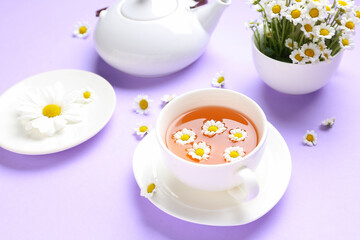 Teapot with cup of natural chamomile tea and flowers on lilac background