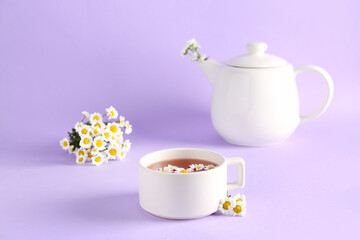 Teapot with cup of natural chamomile tea and flowers on lilac background