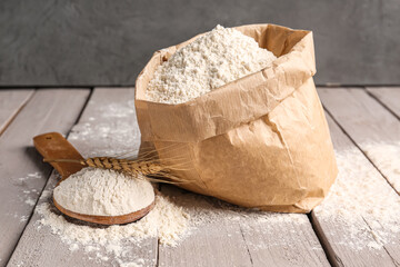 Paper bag with flour, wooden spoon and wheat ear on grey wooden table near wall