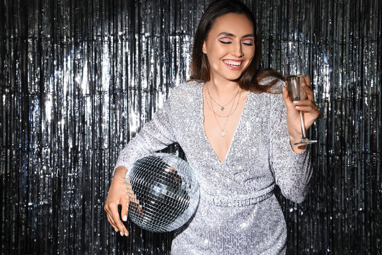 Smiling fashionable woman in sparkling dress with disco ball and glass of champagne against silver tinsel