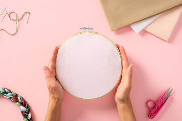 Embroidery concept. Female hands holding round wooden frame with blank canvas over pink background with needlework accessories.