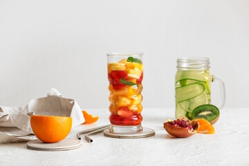 Glass and mason jar of infused water with different sliced fruits on white table