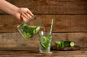Woman pouring infused water with cucumber slices from jug into glass on wooden table