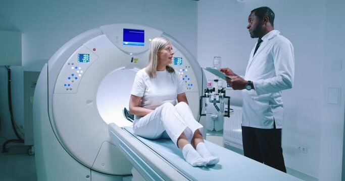 Male doctor and female patient are having discussion at modern tomography laboratory. African American doctor is holding folder and gesturing. Woman sits at MRI bed and talk to doctor.