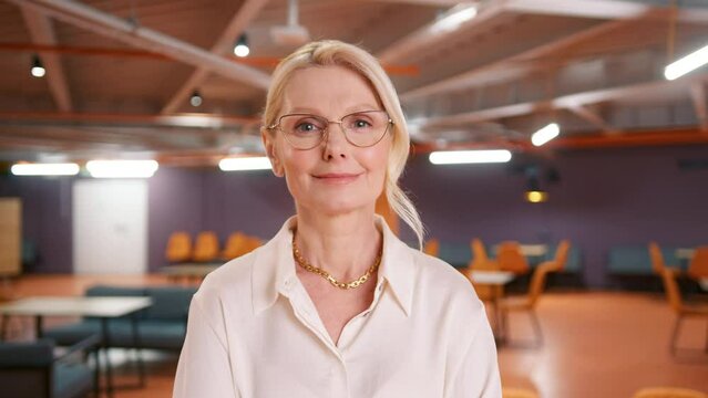 Beautiful smiling confident mature caucasian woman 50s in elegant eyeglasses positive face looking at camera posing alone in empty office, happy professional businesswoman close up front portrait slow
