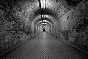 Grayscale of a tunnel with old, dirty walls and lamps on the top of the arched ceilings