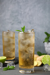 Glass of tasty ginger ale with ice cubes and ingredients on grey wooden table