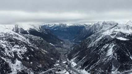 Obraz premium Scenic shot of a village between the snowy mountains covered in evergreen forests on a misty day