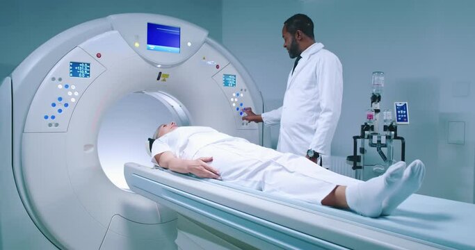 Shoot from side. African American doctor pushes buttons and adjusts MRI capsule for tomography examining. Female patient is lying at CT scanner bed and prepares for magnetic resonance imaging.
