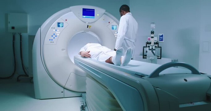 Patient and doctor during magnetic resonance imaging. African American doctor pushes buttons and performs MRI examination. Patient is lying at CT scanner bed amd moving into MRI capsule.