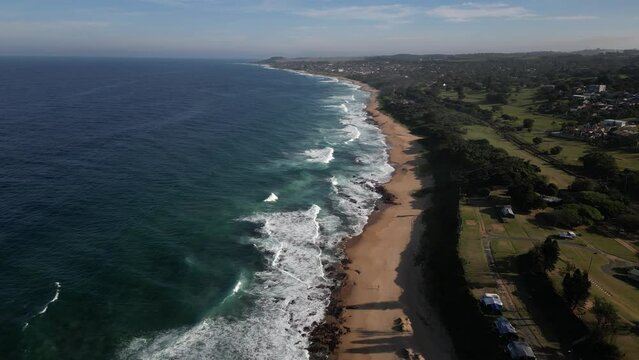 Drone shot over the seaside waves in Scottburgh city with green fields and blue sky, South Africa