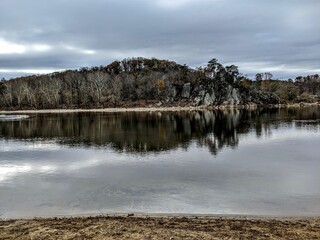 Rocks and the naked trees of the dense forest reflected in the waters of a lake under the gloomy sky