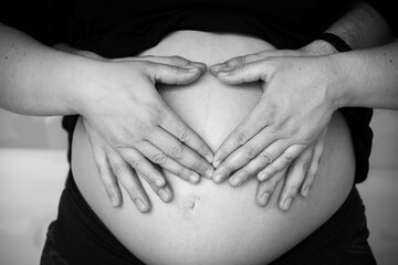close-up shot of a pregnant woman's belly, her and her husband's hands make a heart shape on the...