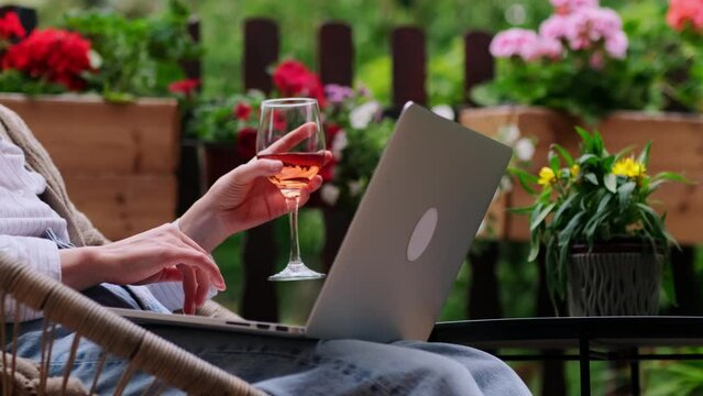Close-up Woman's hand holding a glass of rose wine while using laptop on the cozy home terrace among flowers. Female's fingers slide over the touchpad. Home office, remote work, online communication.