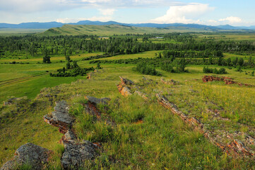 Layered stone formations on top of a high hill overlooking a picturesque valley under a cloudy summer sky.
