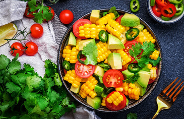 Vegan salad with sweet corn, avocado, jalapeno pepper, red tomatoes, fresh cilantro and olive oil and lime dressing. Black table background, top view
