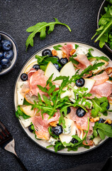Gourmet salad with sweet pears, blueberries, blue cheese, smoked pork ham? arugula and walnuts. Black table background, top view