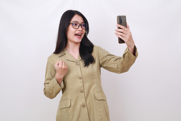 Excited young Indonesian civil servant woman holding a cell phone with fist hand. Isolated on white