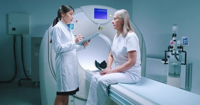 Doctor and patient talk at background of tomography equipment, Female doctor is holding folder and expalining procedure to petient. Patient sit at MRI moving bed and listen to doctor.