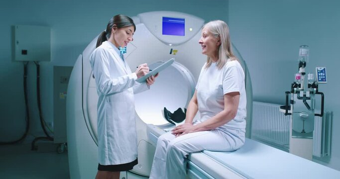 Patient and doctor are talking in tomographic examination laboratory. Doctor holds folder and prescribes treatment to patient. Female patient sits on MRI bed and listens to doctor.