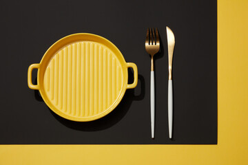Top view of yellow pan with knife and fork displayed on black and yellow background. Abstract presentation for product advertising. Minimal concept, idea for table setting