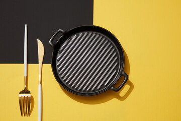 An empty black pan placed on black and yellow background with set of knife and fork. View from...
