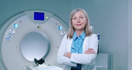 Portrait of female doctor at background of MRI capsule. Middle-aged doctor dressed up in medical uniform is posing for camera. Woman doctor with arms crossed is looking straight at camera.
