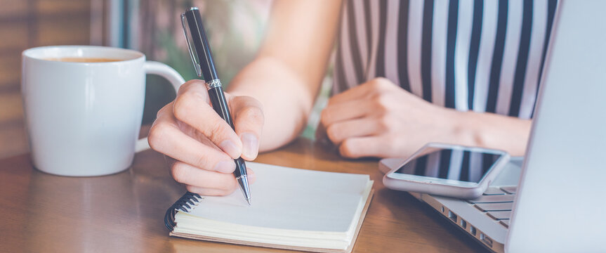 Woman hand is writing on a notepad with a pen on a wooden desk.