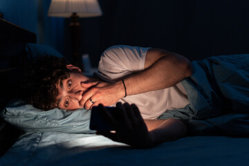 Shocked young man looking at mobile phone in bed at home at night, insomnia and addiction