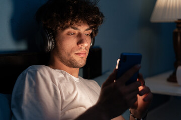 Portrait of a serious guy listening to music with headphones and smart phone lying on the bed in the night at home, watching movie online