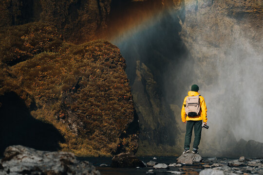 Man in yellow raincoat photographs waterfall with rainbow overhead