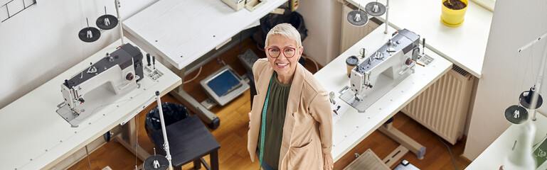 Obraz premium Middle aged woman designer looks at camera standing near sewing machines in spacious studio