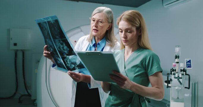 Female doctor and nurse standing at equipped tomography room. Doctor is holding scans and consulting with colleague. Nurse hold folder and listen to doctor carefully. Analizing of examination results,