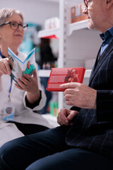 Senior woman apothecary specialist giving customer pharmaceutical consultancy service, reading patient medical brochure about elderly healthy lifestyle. Old man holding heart supplements in drugstore