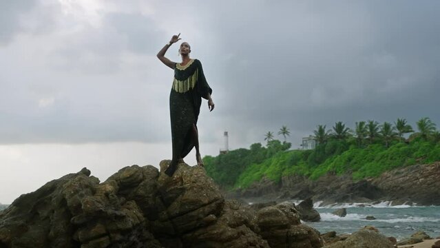 Divine non-binary black person in boutique dress, brass jewelry poses on rocky hill top with dramatic ocean cloudy skyline. Queer lgbtq fashion model in posh outfit fluttering in air. Pantheon concept