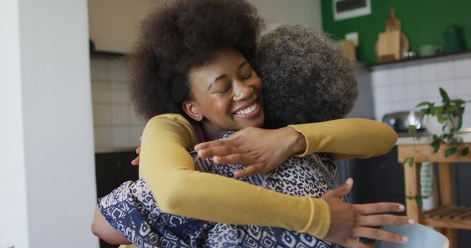 Happy african american adult daughter and senior mother hugging in kitchen at home, slow motion
