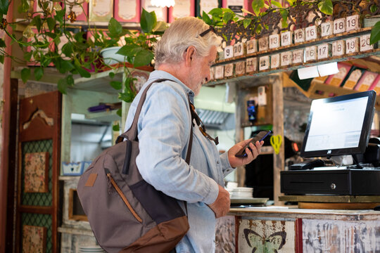 Senior white-haired man pays at the coffee shop checkout using the application on his mobile phone. Concept of modernity and online electronic payments