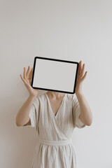 Tablet in female hands. Woman in neutral tan beige linen dress over white wall. Boho, bohemian fashion style. Aesthetic mock up with empty free copy space blank pad screen