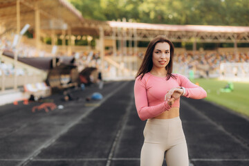 Fototapeta premium Portrait of athletic attractive woman in sportswear standing at the football stadium and looking on the indicators on her sport watch, healthy lifestyle.