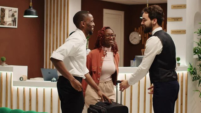 Happy african american tourists tipping helpful bellboy carrying their luggage in resort lobby. Cheerful guests checking out from hotel after handing over room access key card to friendly receptionist