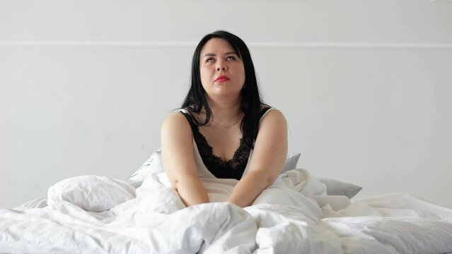 Black-haired lady getting up with weary and powerless expression from cozy bed. Plump woman with exhausted expression has trouble sleeping at night