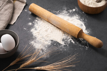 Organic ingredients for baking, scattered wheat flour, eggs, rolling pin and kitchen textiles on a dark gray  background.