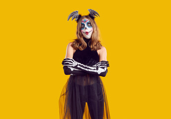 Studio portrait of child in Halloween costume. Teenage girl with sugar skull make up standing with arms folded isolated on yellow background and looking at camera with funny surprised face expression