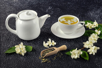 Green tea with jasmine in a white cup and teapot on a dark background. Fresh jasmine flowers and a wooden spoon as decor